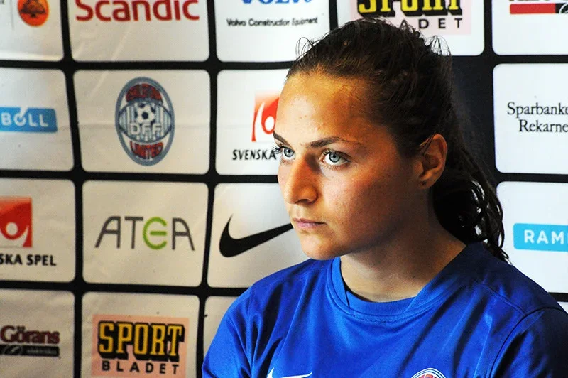 Elena Sadiku me bluzë sportive blu, përpara një muri me logot e sponsorëve.